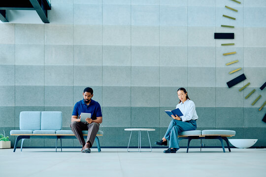 Two diverse business people waiting in a contemporary office lobby, with one using a digital tablet and another reading a book