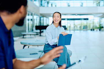 Fototapeta premium Businesswoman holding a notebook, smiling and discussing a project with a colleague in a modern office lounge area