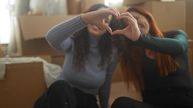 Woman forming heart with hands while sitting among moving cardboard boxes in apartment building; friendship joy.