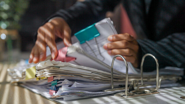 Hands organize papers with focused care. Papers stack high, clipped with colorful tabs. Binder rings gleam under soft desk light. Person's intent shows dedication to tasks. Ideal for business, office