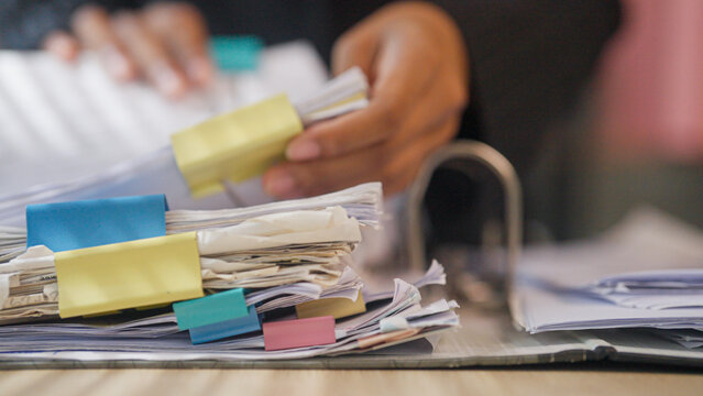 Hands sort papers stacked with colorful clips. Desk clutter suggests busy work or urgent tasks. Focused action implies organization or decision-making. Ideal for business, office