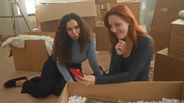 Two women friends unpacking cardboard boxes, hand holding red smartphone, kneeling and pointing at screen inside building; supportive friendship.