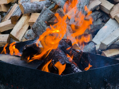 Bright orange tongues of flame burning wood inside a black metal brazier or grill against a background of stacked firewood billets.