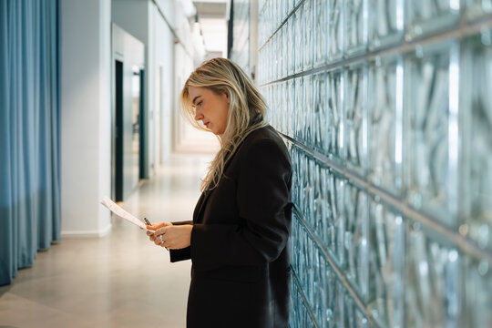 Side view of businesswoman reading reports while leaning on glass brick wall at office