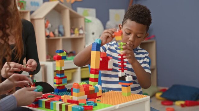 Boy stacks colorful blocks with hand while woman helps build a tower at table in building; creative learning concentration.