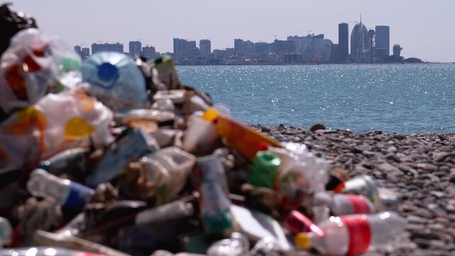 Large pile of plastic bottles and other non-degradable waste contaminating a pebble beach, with the glittering sea and a distant urban skyline creating a stark contrast