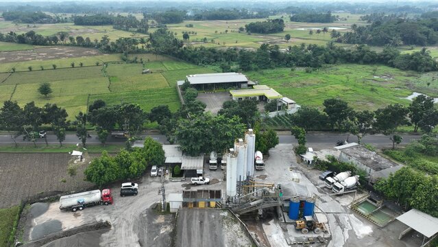 Cianjur Regency, Indonesia - 13 March 2025: Aerial view of a concrete batching plant with silos and mixer trucks surrounded by lush green rice fields in West Java.