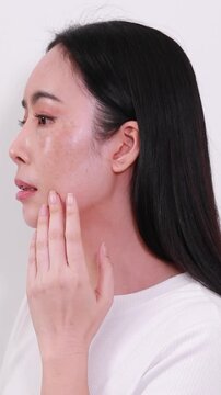 Side view of an Asian woman looking worried while touching her face, showing visible melasma, freckles, and dark spots on her cheek against a plain background.
