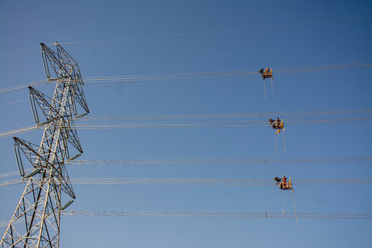 Joypurhat, Bangladesh - 12 November 2024: Aerial view of electrical workers in maintenance baskets suspended from high voltage power lines against a clear blue sky.
