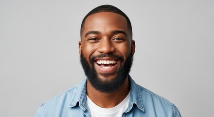 Obraz premium Portrait of a Happy Young African American Man with a Beard Laughing and Smiling in a Denim Shirt on a Gray Background