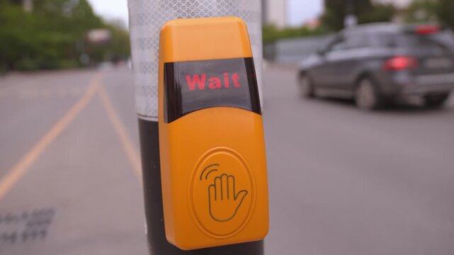 Closeup of a person's hand pressing a yellow pedestrian call button at a crosswalk to activate the traffic signal, with cars passing in the background on a city street