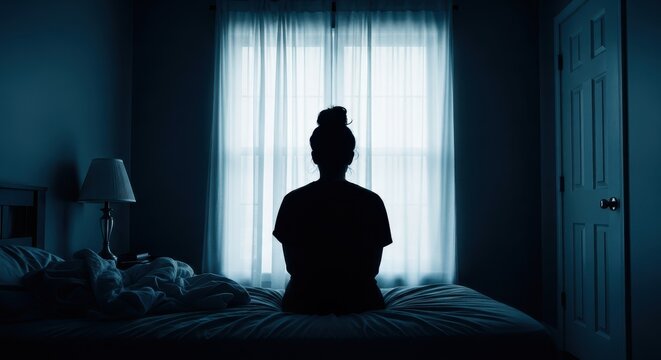 Silhouette of a lonely woman sitting on a bed in a dark room facing a window, concept of depression and mental health