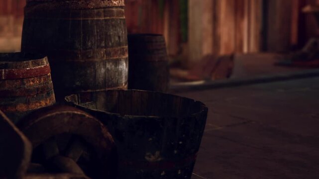Wooden barrels and containers are placed on the floor of a rustic indoor space. The area has warm light, and wooden elements are visible. People can be seen in the background.