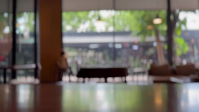 Blurred modern coffee shop interior with wooden tabletop foreground, large glass windows, soft natural daylight, warm pendant lighting, cozy seating area and spacious copy space for advertising.
