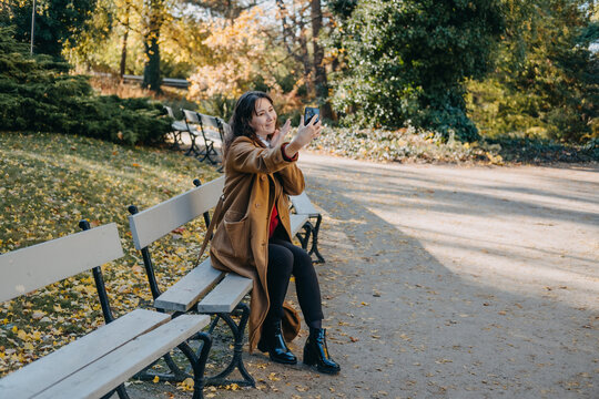 Woman sits on bench in public park while taking selfie photo with smartphone. Influencer social media campaign, lifestyle blogger recording outdoor, digital storytelling visual asset.