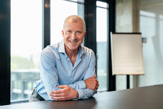 Smiling business executive leaning on desk in modern office