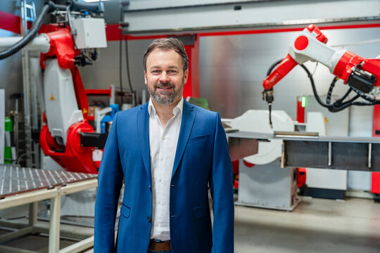 Smiling manager standing indoors near industrial robot in factory