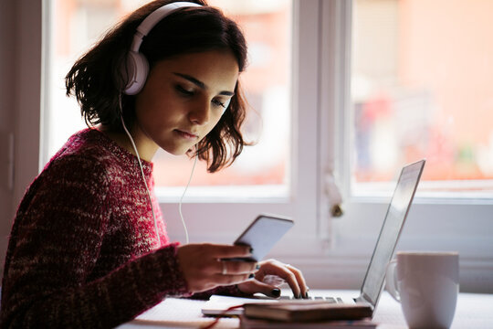 Young woman with laptop and headphones looking at cell phone