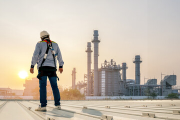 Industrial Sunrise: A lone worker, outfitted with safety gear, stands on a rooftop, surveying a vast industrial complex as the sun rises, casting a warm golden hue over the scene. © Rassarin
