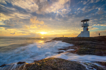 Lighthouse Sentinel at Sunrise: A solitary lighthouse stands guard over a rocky coastline as the sun breaks the horizon, casting a golden light over the churning ocean waves. © Rassarin