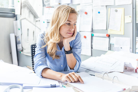 Pensive woman sitting at desk in office surrounded by paperwork