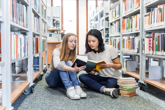 Two teenage girls sitting on the floor in a public library reading in a book