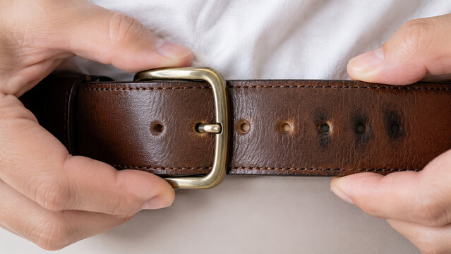 Close-up of hands buckling a leather belt at a notch smaller than the worn notches beside it. Weight loss progress concept metaphor.