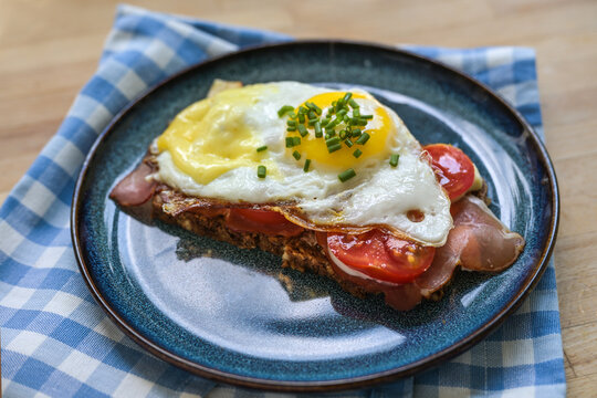 Dark whole-grain bread sandwich with ham, tomatoes and a fried egg, topped with melted cheese and chives on a blue plate, breakfast or light lunch, selected focus