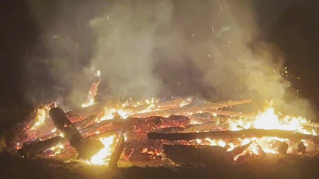 Large midsummer bonfire burning outdoors at night. Flames, glowing embers, and smoke rise into the dark sky.