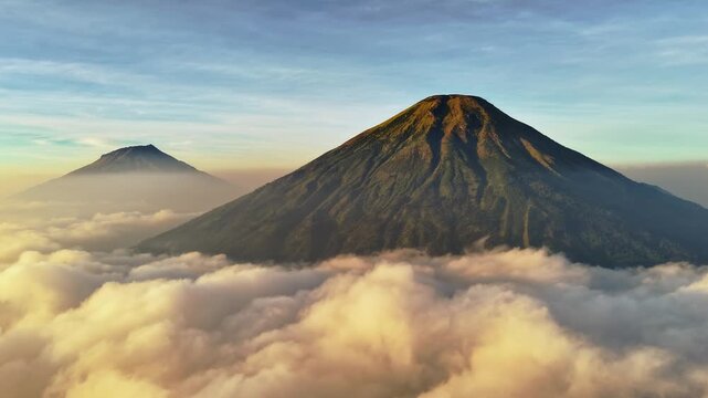 Aerial view of Mount Sindoro and Mount Sumbing rising above a sea of clouds during golden hour in Kecamatan Kedu, Jawa Tengah, Indonesia.