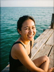 Fototapeta premium Young woman laughing while sitting on a wooden pier by the sea with warm sunlight. The subject conveys joy and relaxation near water in a casual summer setting.