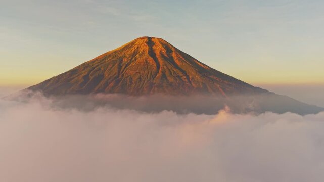 Aerial view of Mount Sindoro peak rising above a thick layer of morning clouds with golden sunlight hitting the volcanic slopes in Kecamatan Kedu, Jawa Tengah, Indonesia.