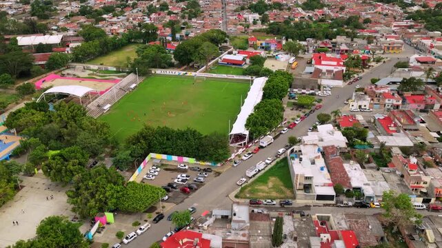 Estadio Municipal de Tuxpan with a green green field, covered stands, surrounding trees, streets, parked cars, and nearby neighborhoods near Tuxpan, Jalisco. Aerial drone view