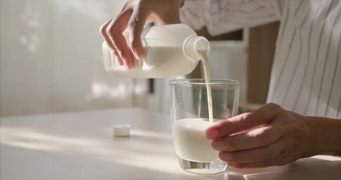 Close up of Asian Woman Hands Pouring Soy Milk into Glass on Table in Cozy Living Room with Warm Morning Sunlight.