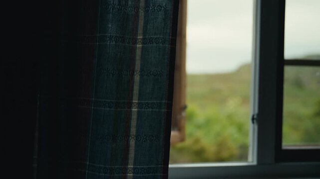 A dark woven curtain hangs beside an open window, where soft daylight and a distant mountain view create a quiet interior scene filled with stillness, nostalgia, and old rural character.