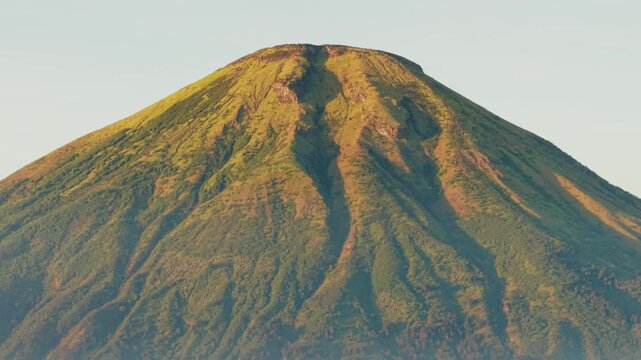 Aerial view of the lush green slopes and volcanic peak of Mount Sindoro under soft morning light in Kecamatan Kedu, Jawa Tengah, Indonesia.