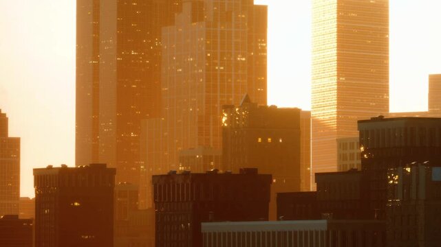 Sunlight reflects off skyscrapers as it sets in the evening. The buildings create a striking city skyline with different shapes and heights against a warm glow.