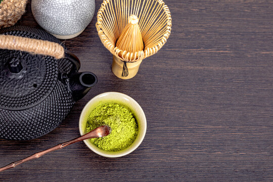 Matcha green tea powder in small bowl with bamboo whisk and cast iron teapot on wooden surface. Traditional Japanese tea ceremony equipment for healthy organic hot beverage preparation