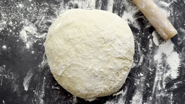 Dough ball on a black countertop with flour scattered around, a wooden rolling pin positioned nearby, ready for rolling out into a flat shape for baking