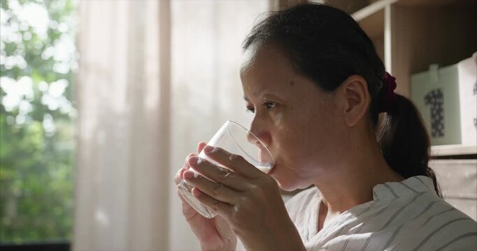 Close up face. Asian Woman Drinking Soy Milk from Glass in Living Room, Female Enjoying Plant-Based Milk Alternative by Window with Morning Sunlight and Curtains, Healthy Lifestyle Routine.