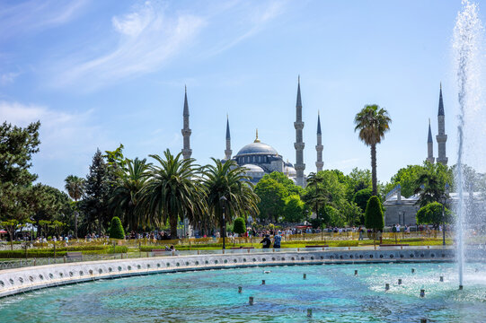 The iconic Sultan Ahmed Mosque with six minarets is seen across a sparkling park fountain and palm trees on a sunny summer day in Istanbul's Sultanahmet district.
