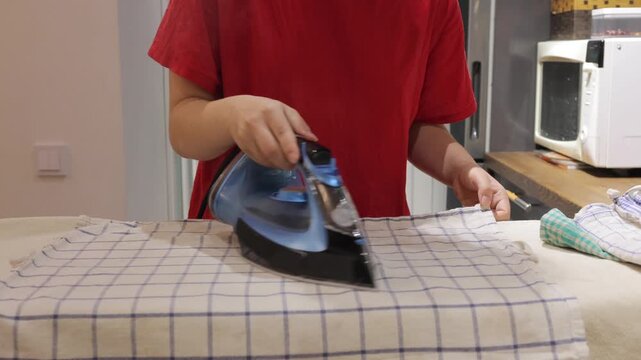 Hands guide a steam iron over a checked dishcloth on an ironing board in an indoor kitchen.