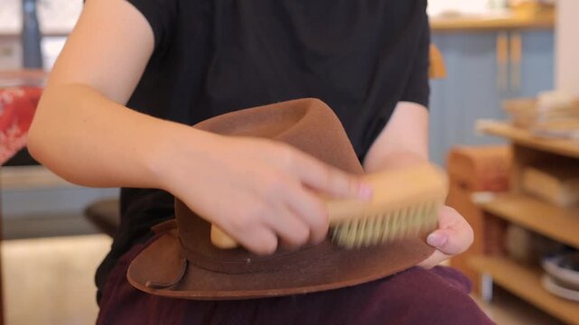 Hands clean a brown wide brim hat with a wooden brush in a soft indoor close-up about careful clothing maintenance.