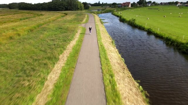 Aerial view of a person jogging along a paved path beside a canal in the green countryside of Berkel en Rodenrijs, South Holland, Netherlands.