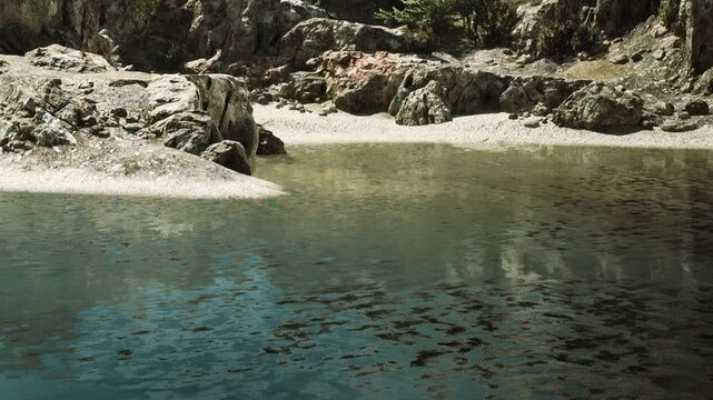 Rocks and shallow water are seen at a natural lake in a remote area. The sunlight reflects off the water surface. Trees line the background, making the setting appear more isolated.