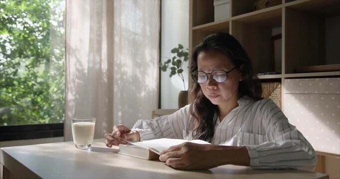 Side view. Middle Aged Asian Woman with Glasses Reading Book in Living Room, Asia women Soy Milk on Table by Window in Warm Morning Sunlight, Cozy Home Interior.