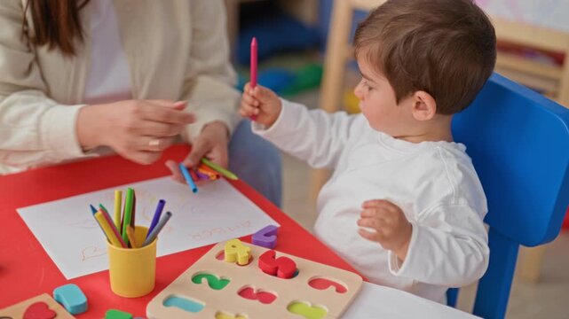Woman offering crayons to little boy at preschool table with wooden number puzzle, paper and cup of colored pencils, hands visible; calm learning.
