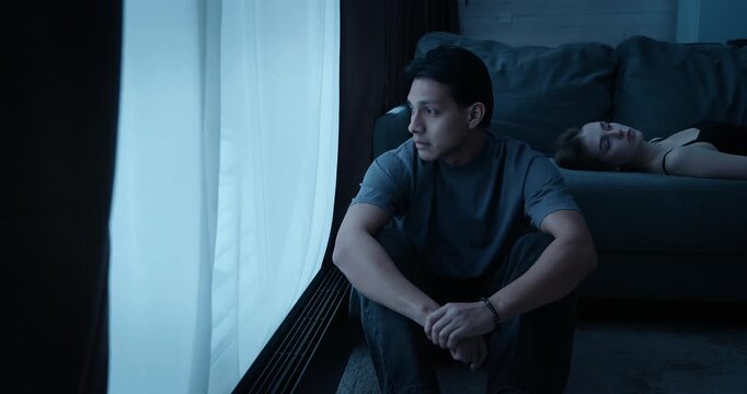 Depressed man sit window looking out, woman lie sofa dark room at home. Couple quarrel ignoring problem. Relationship crisis, misunderstanding, loneliness, sadness concept. Family conflict brake up.