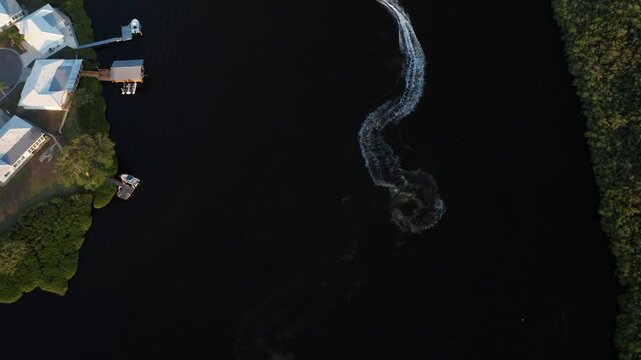 Riverside houses and docks border a dark channel as a fast boat cuts a curving foam trail between wooded banks at dusk.