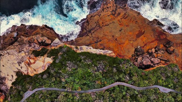 Aerial view of rugged sandstone cliffs with crashing ocean waves, dense green vegetation, and a winding coastal road in New South Wales, Australia.